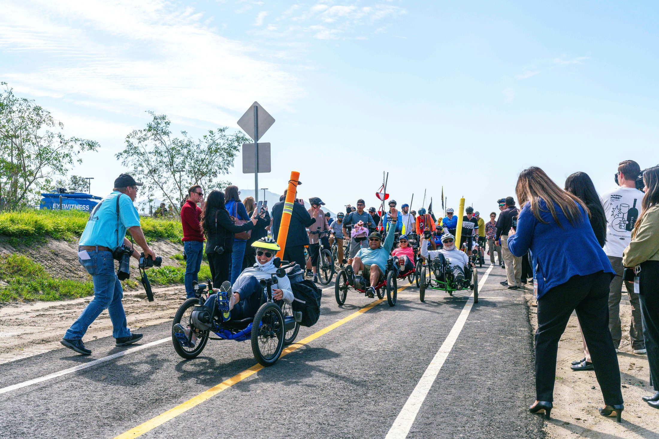 A group of trike cyclists ride in a line flanked by onlookers taking photos on the Santa Ana River Trail in San Bernardino