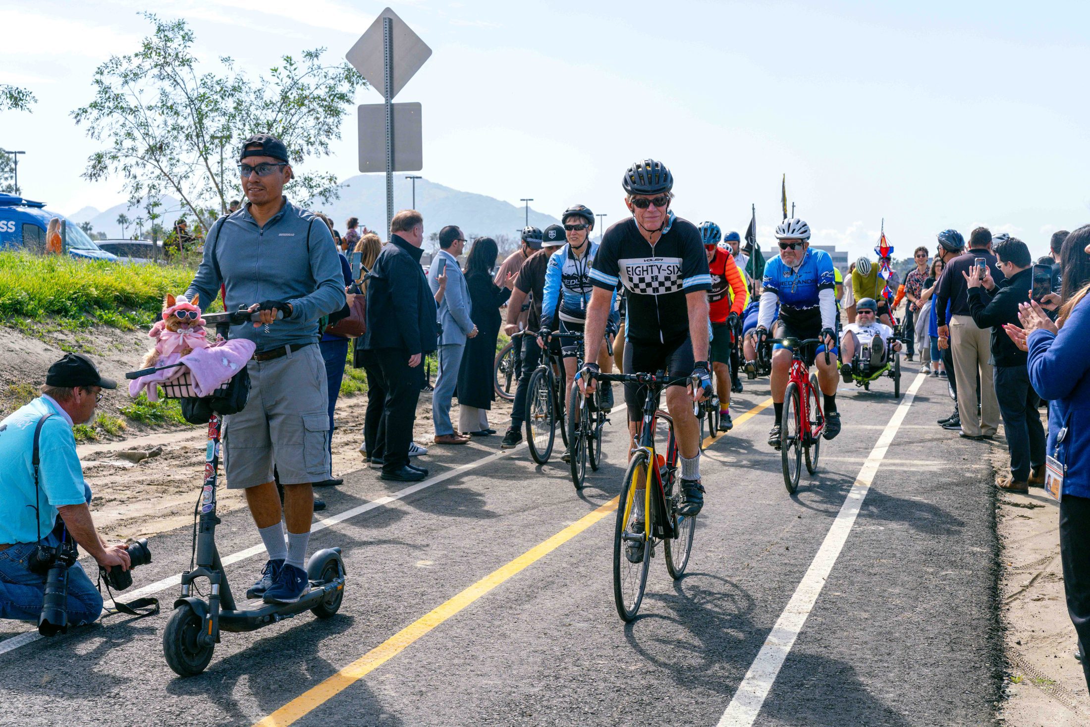 Three bicyclists and a man on a scooter with a small dog dressed in pink in a front basket ride in a line on the Santa Ana River Trail.