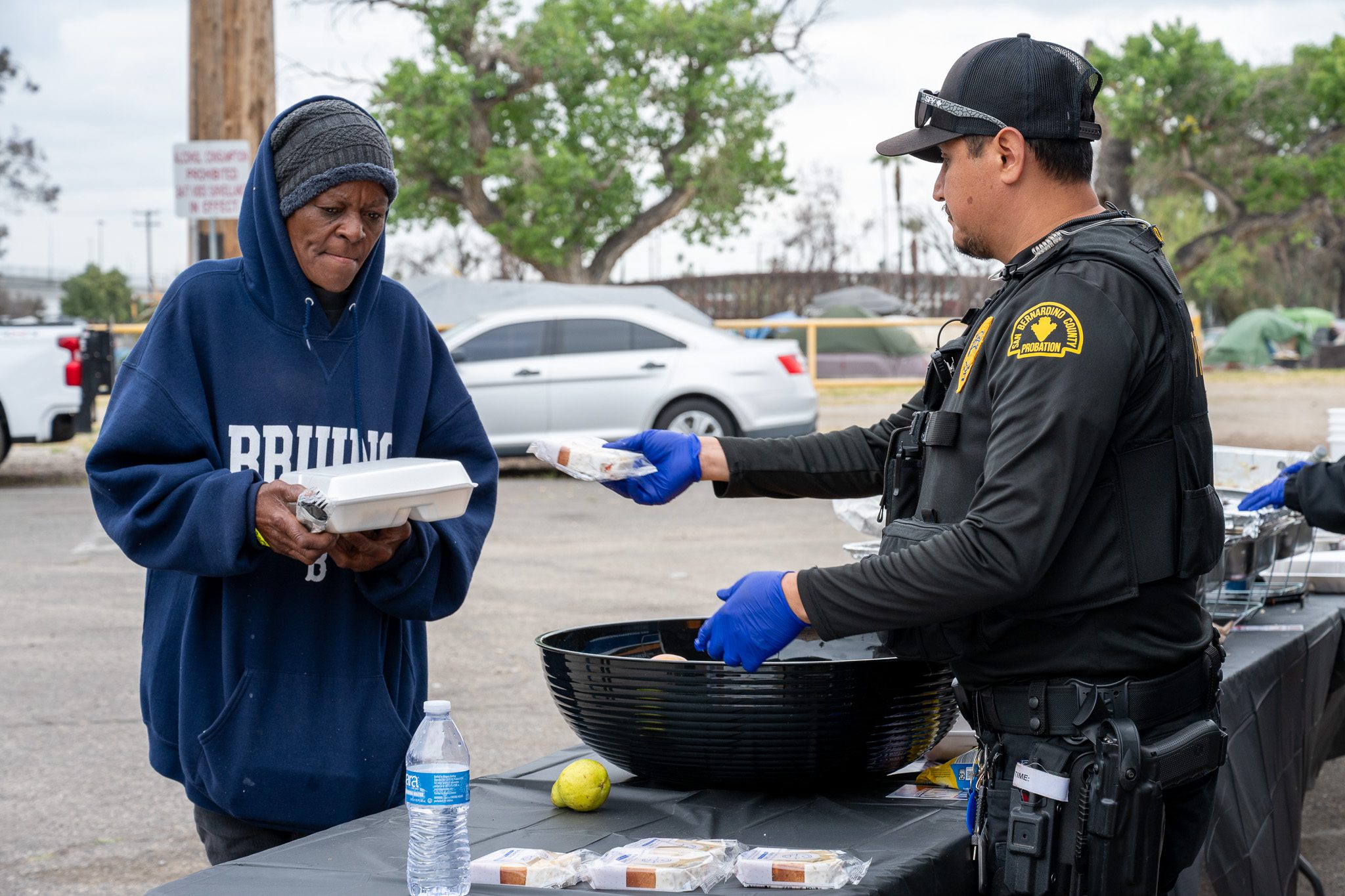 A woman in a blue hoodie holds a meal box while an SBC Probation officer offers her a packaged meal beside a fruit bowl.