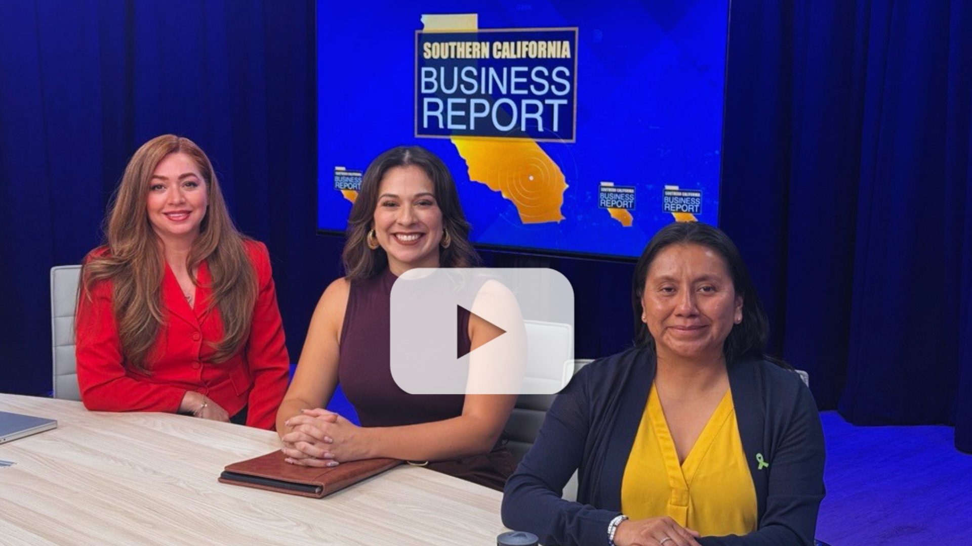 Three women sit at a table in a TV studio with a Southern California Business Report sign on a screen behind them, with a video play button overlay indicating a video.