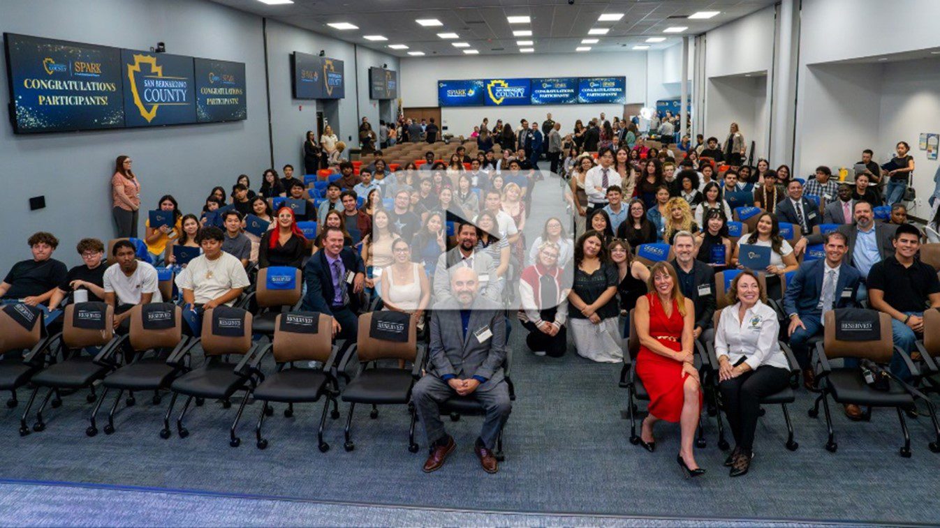 photo of SPARK program participants, county leaders and staff posing for a photo in a conference room with a video play button overlay.