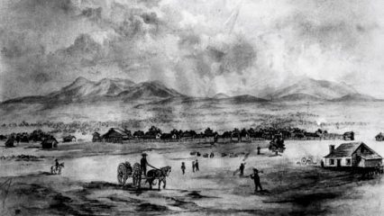 Black and white photo featuring a small town with houses, settlers and a horse-drawn cart with mountains in the background.