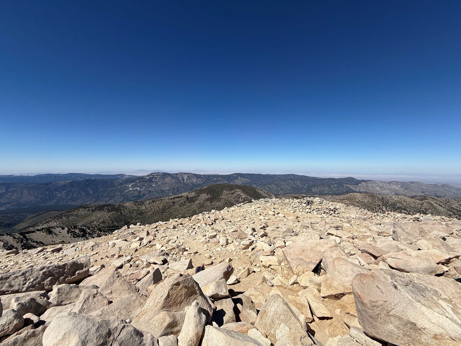A view from the rocky summit of San Gorgonio Peak, showcasing an expansive landscape of distant mountains and a clear blue sky.