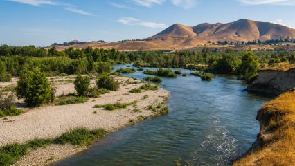 A landscape featuring a gently flowing river bordered by green and sandy banks. In the background, rolling hills contrast with a blue sky dotted with wispy clouds.