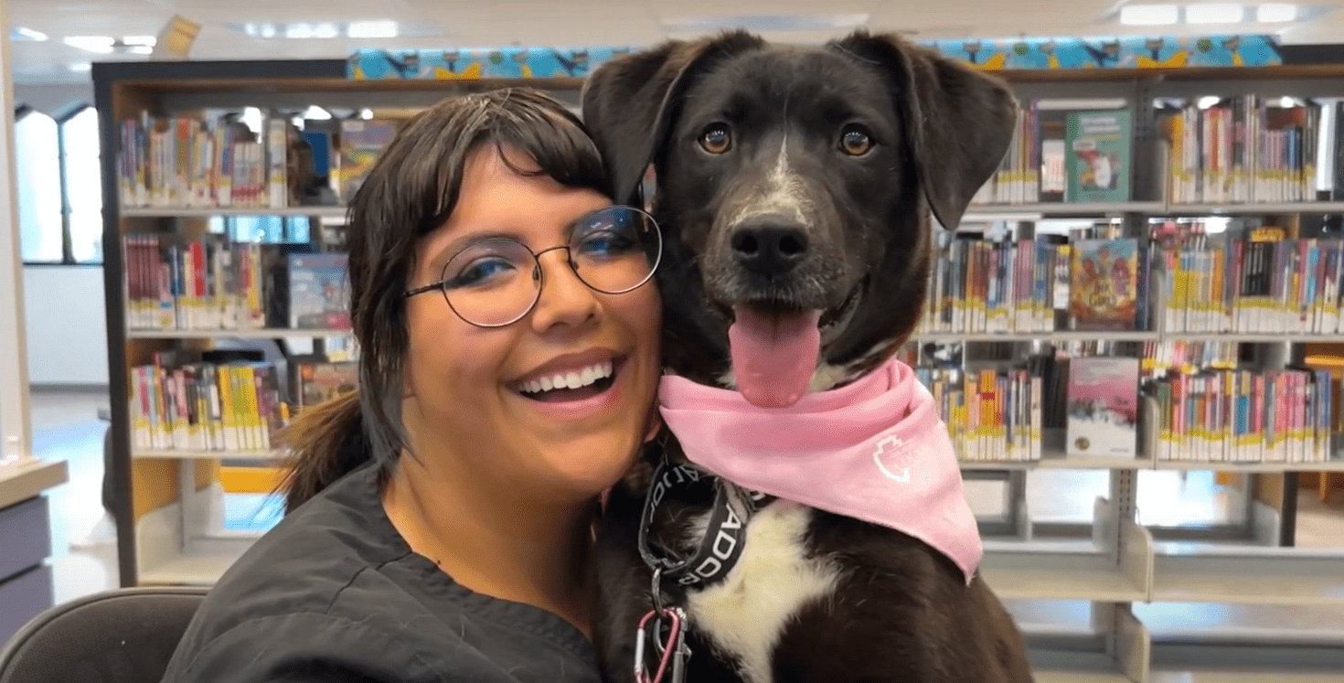 Play button, staff member hugging a black and white dog with a scarf on inside a library, San Bernardino County log.