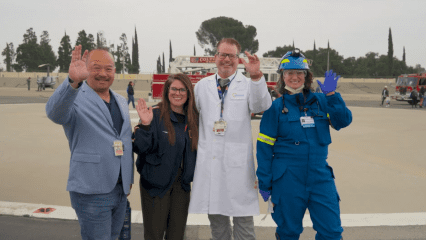 Four emergency and medical professionals—two women and two men—stand side by side on a helipad, smiling and waving at the camera. They are dressed in various uniforms including a suit jacket, paramedic gear, a white lab coat, and full emergency response attire. In the background are fire trucks, a helicopter, and several other responders.