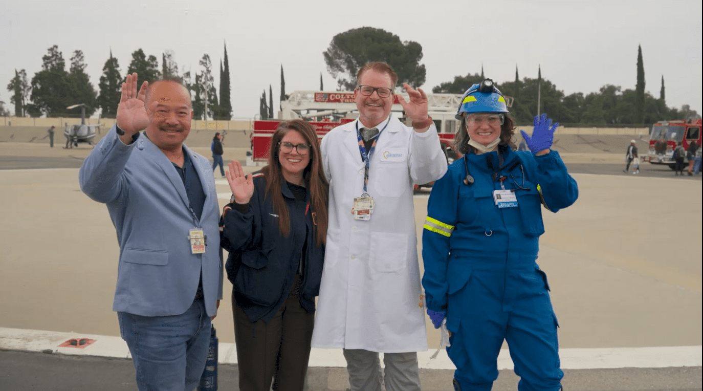 Four emergency and medical professionals—two women and two men—stand side by side on a helipad, smiling and waving at the camera. They are dressed in various uniforms including a suit jacket, paramedic gear, a white lab coat, and full emergency response attire. In the background are fire trucks, a helicopter, and several other responders.