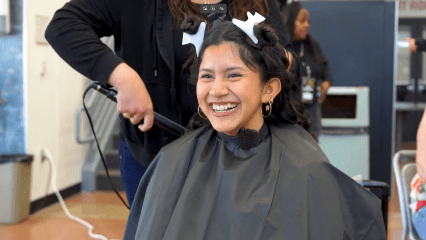 A smiling young woman with hair clips sits in a salon chair, wearing a black cape. A stylist in black is curling her hair, creating a joyful atmosphere.