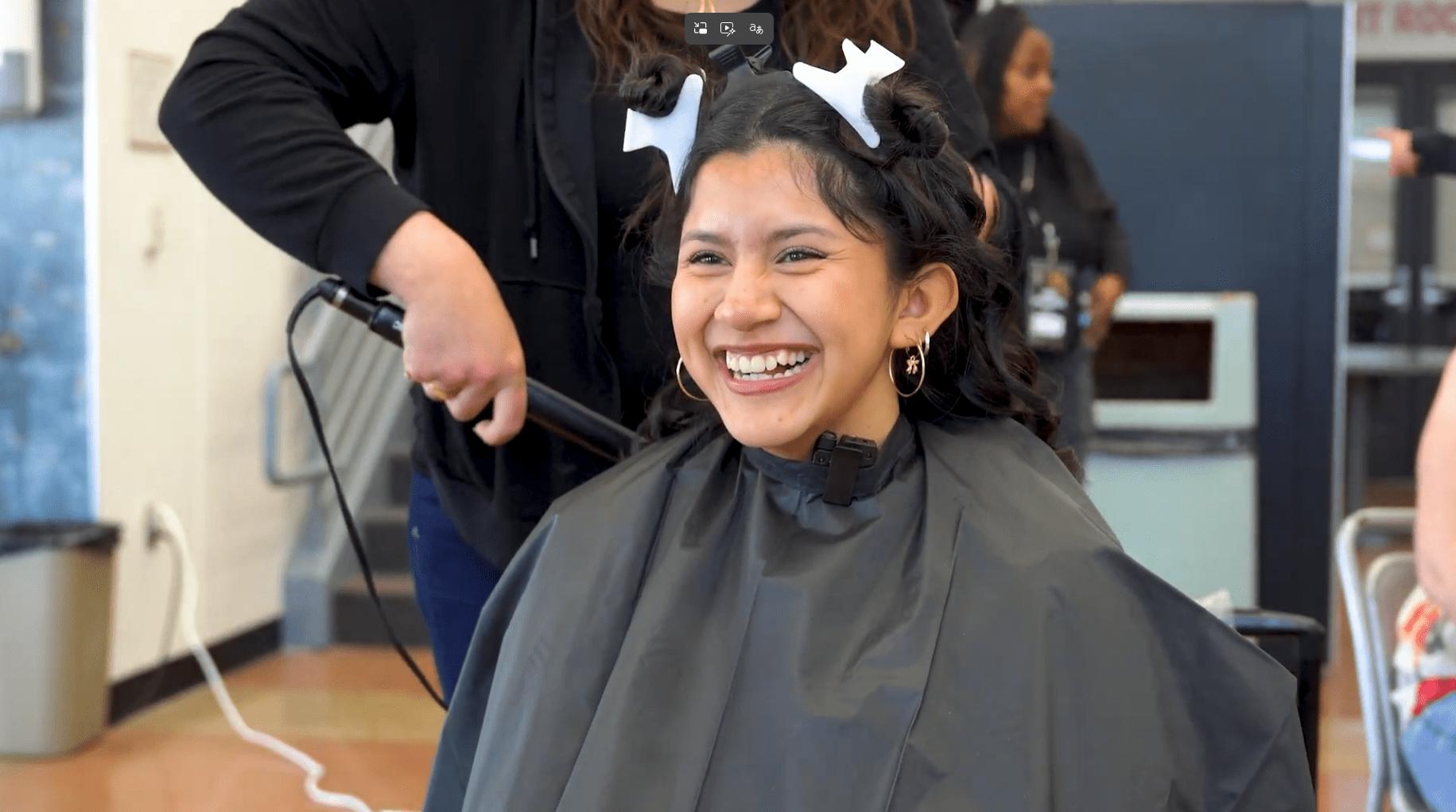 A smiling young woman with hair clips sits in a salon chair, wearing a black cape. A stylist in black is curling her hair, creating a joyful atmosphere.