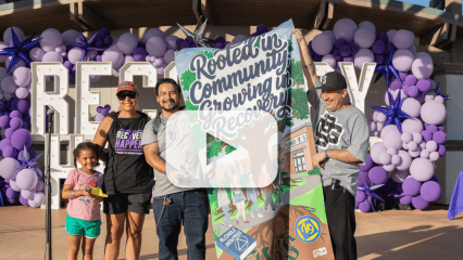 Recovery Happens participants hold a colorful “Rooted in Community, Growing in Recovery” banner in front of a purple balloon display.