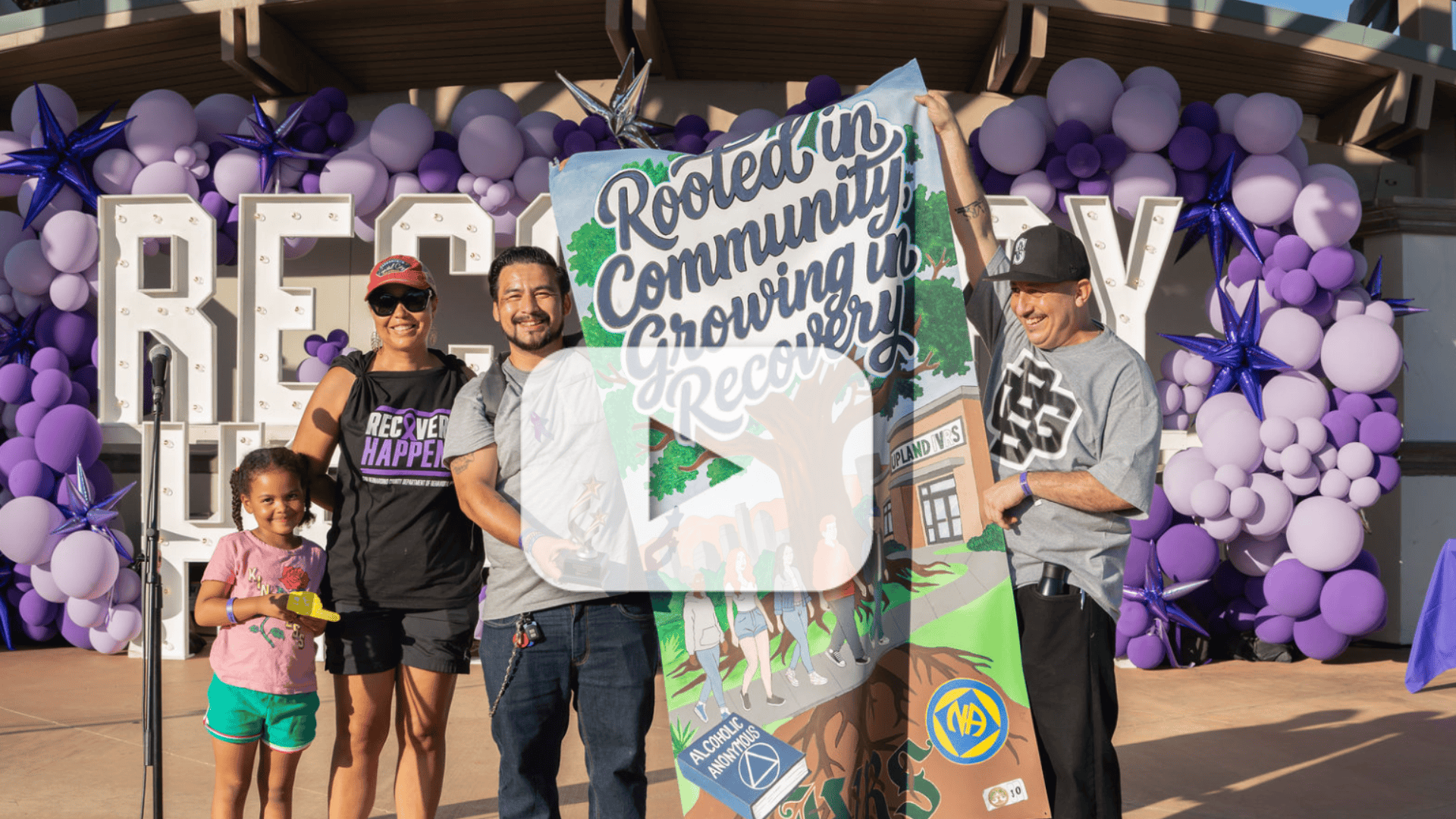 Recovery Happens participants hold a colorful “Rooted in Community, Growing in Recovery” banner in front of a purple balloon display.