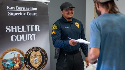 A Probation staff member hands a document to a man. Also visible is a San Bernardino Superior Court Shelter Court sign.