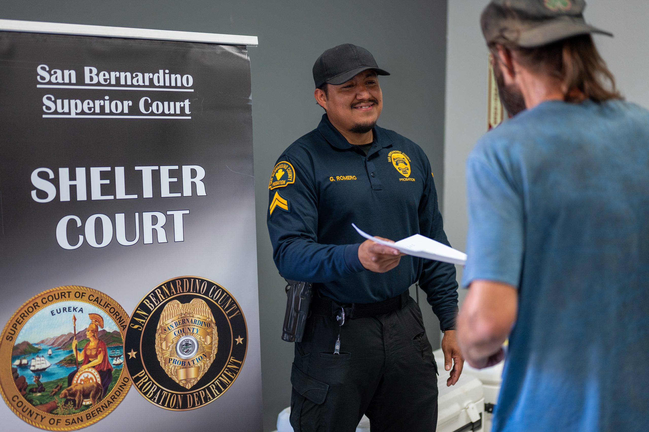 A Probation staff member hands a document to a man. Also visible is a San Bernardino Superior Court Shelter Court sign.