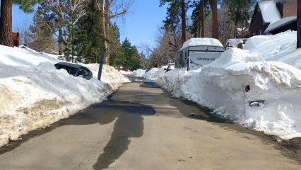 A mountain road cleared down the center for traffic, with snow-covered cars parked along both sides, showing the impact of not moving vehicles before plowing.