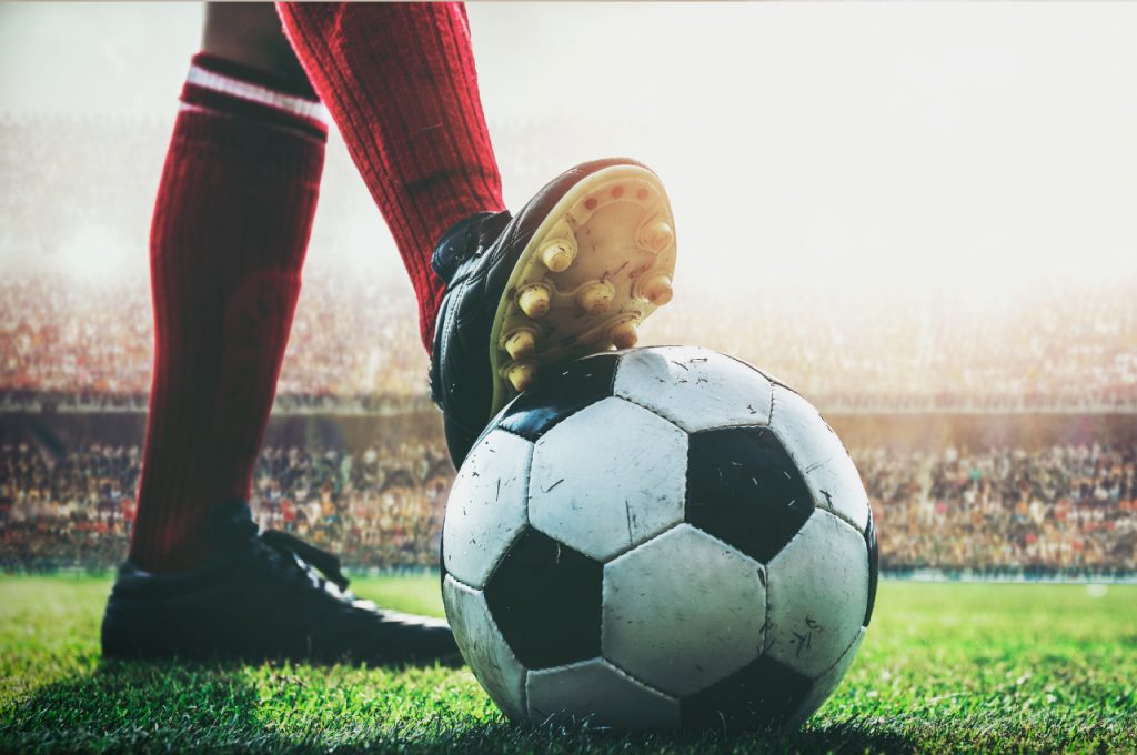 A soccer player’s foot rests on a soccer ball on a field with a large stadium crowd in the background.