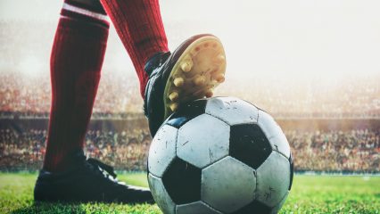 A soccer player’s foot rests on a soccer ball on a field with a large stadium crowd in the background.