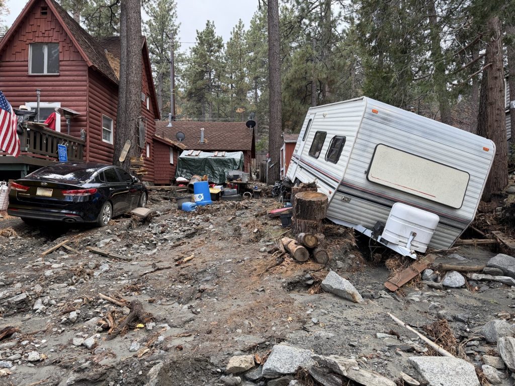 Flood- and debris-damaged mountain home with mud-covered driveway, displaced trailer and damaged vehicles.