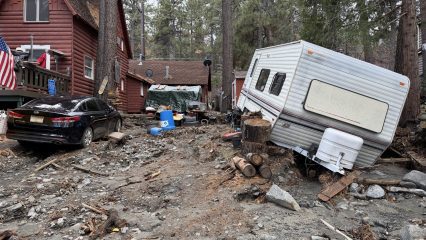 Flood- and debris-damaged mountain home with mud-covered driveway, displaced trailer and damaged vehicles.