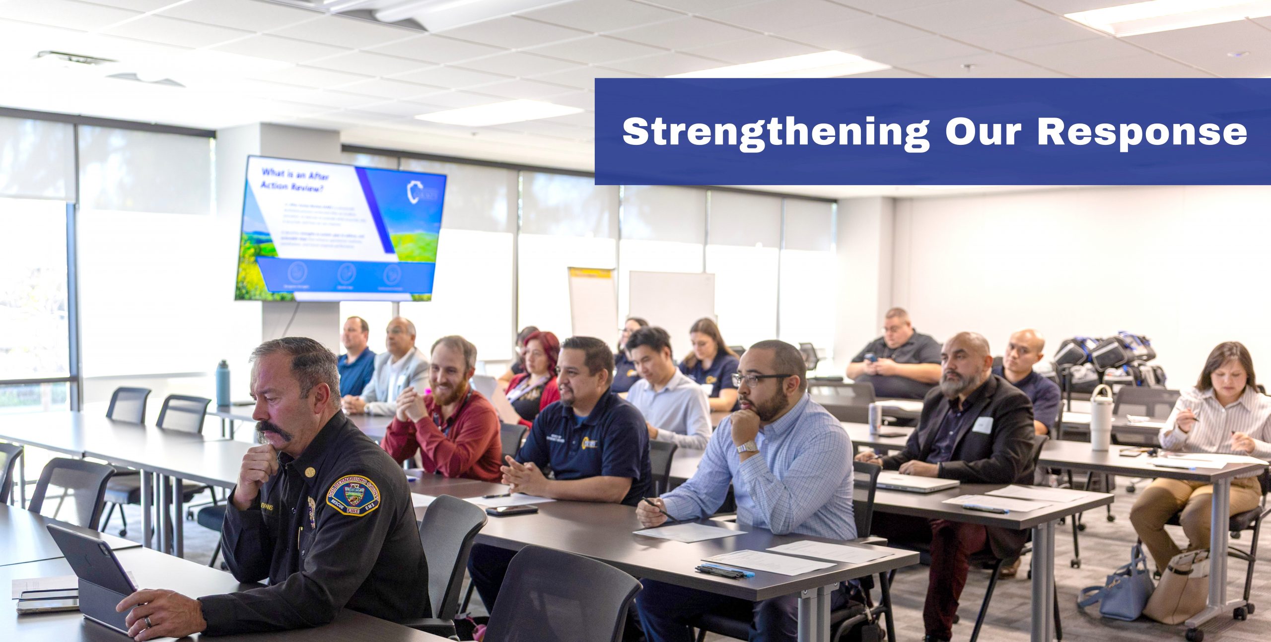 Attendees sit in a conference room during a “Strengthening Our Response” training, viewing a slide on After Action Reviews.