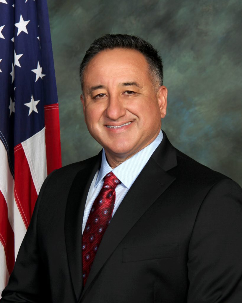 A formal headshot of Supervisor Joe Baca, Jr. in a black suit and red tie with an American flag to his side.