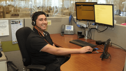 A smiling young male employee in a black shirt, wearing a headset, sitting at a desk in a customer service center.