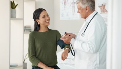 Male doctor checks a smiling female patient's blood pressure during an office visit in a medical exam room. Anatomy charts hang on the wall.