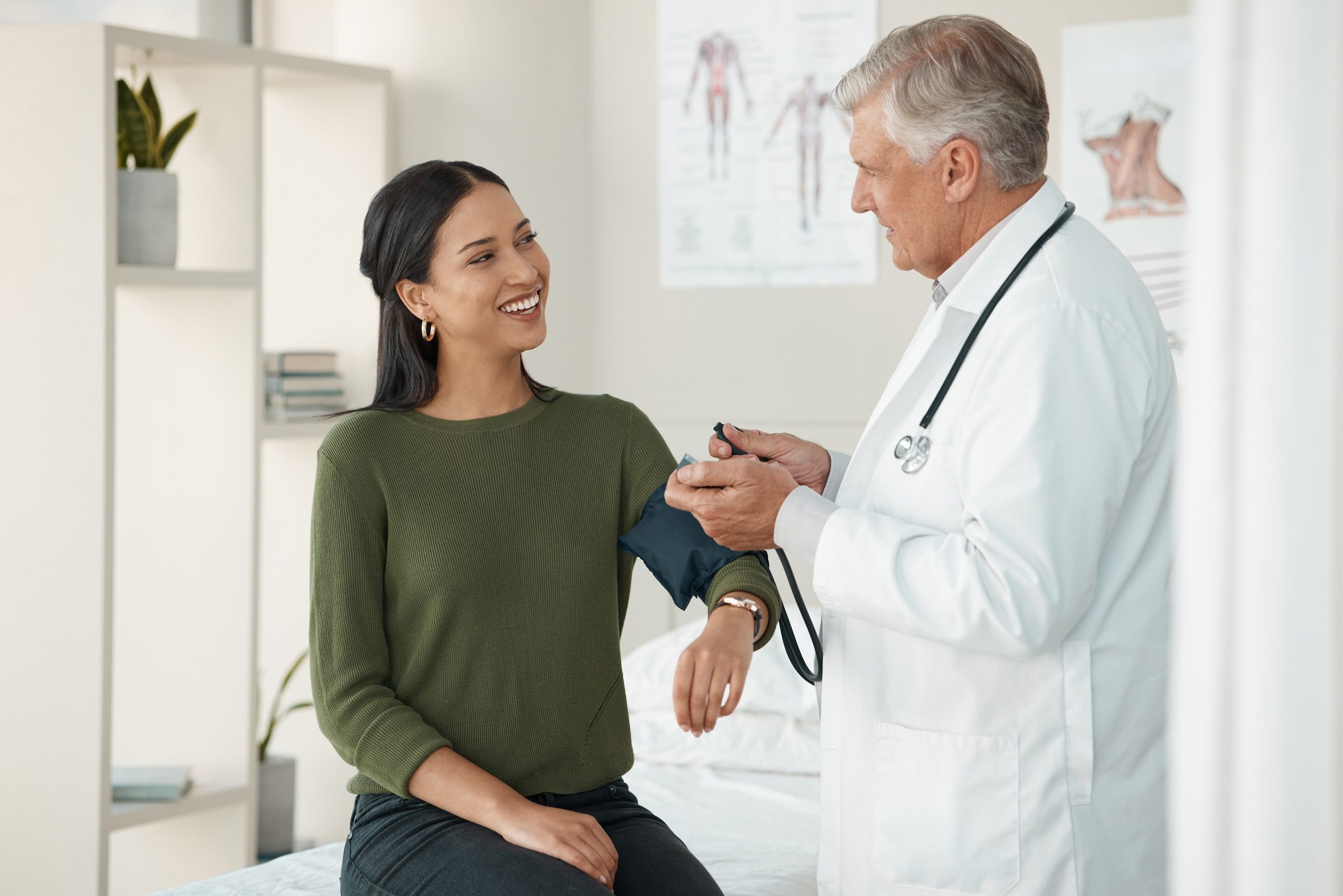Male doctor checks a smiling female patient's blood pressure during an office visit in a medical exam room. Anatomy charts hang on the wall.