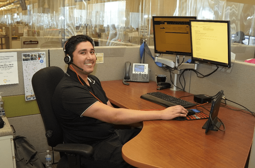 A smiling young male employee in a black shirt, wearing a headset, sitting at a desk in a customer service center.  