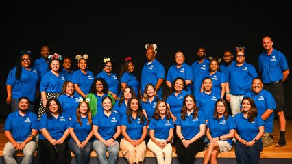 Group photo of Wellness Conference 2025 planning team wearing matching polos and fun headbands, standing on a stage.