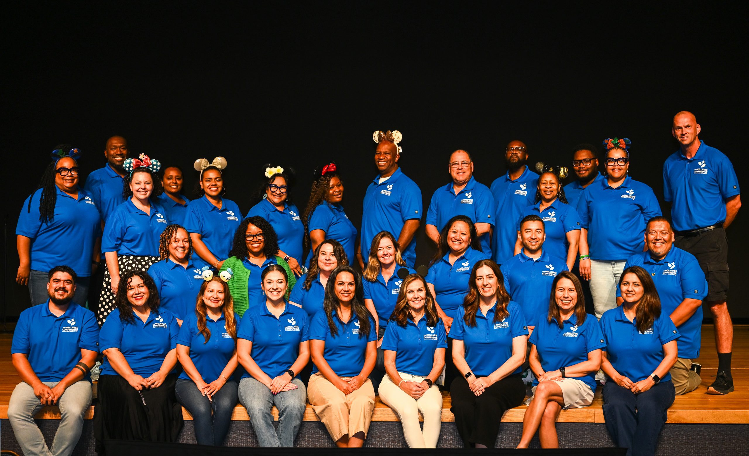 Group photo of Wellness Conference 2025 planning team wearing matching polos and fun headbands, standing on a stage.