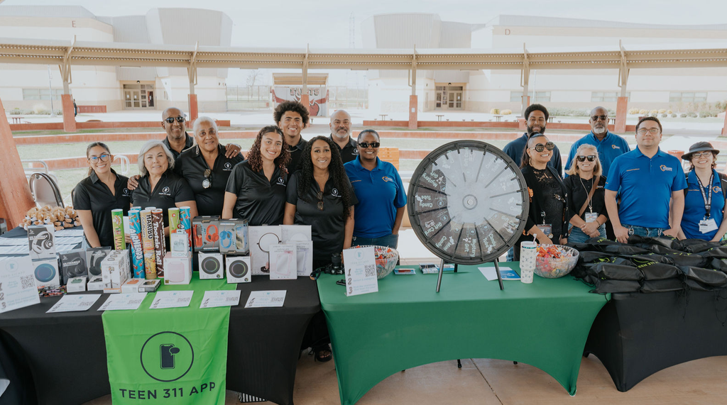 Teen 311 App team members pose behind an outreach table with giveaways, prizes and information at an outdoor community event.