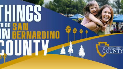 A banner reading “Things to Do in San Bernardino County” featuring a vibrant design with blue and gold accents. On the right, a woman and a laughing child enjoy a sunny day at an outdoor event with tents in the background. The San Bernardino County logo is included, along with stylized tree silhouettes.