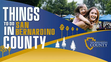 A banner reading 'Things to Do in San Bernardino County' featuring a vibrant design with blue and gold accents. On the right, a woman and a laughing child enjoy a sunny day at an outdoor event with tents in the background. The San Bernardino County logo is included, along with stylized tree silhouettes.