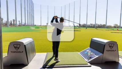 A person is taking a swing on an outdoor driving range at Topgolf. The area features green turf, targets in the distance, and a protective netting surrounding the range, and video play button overlay.