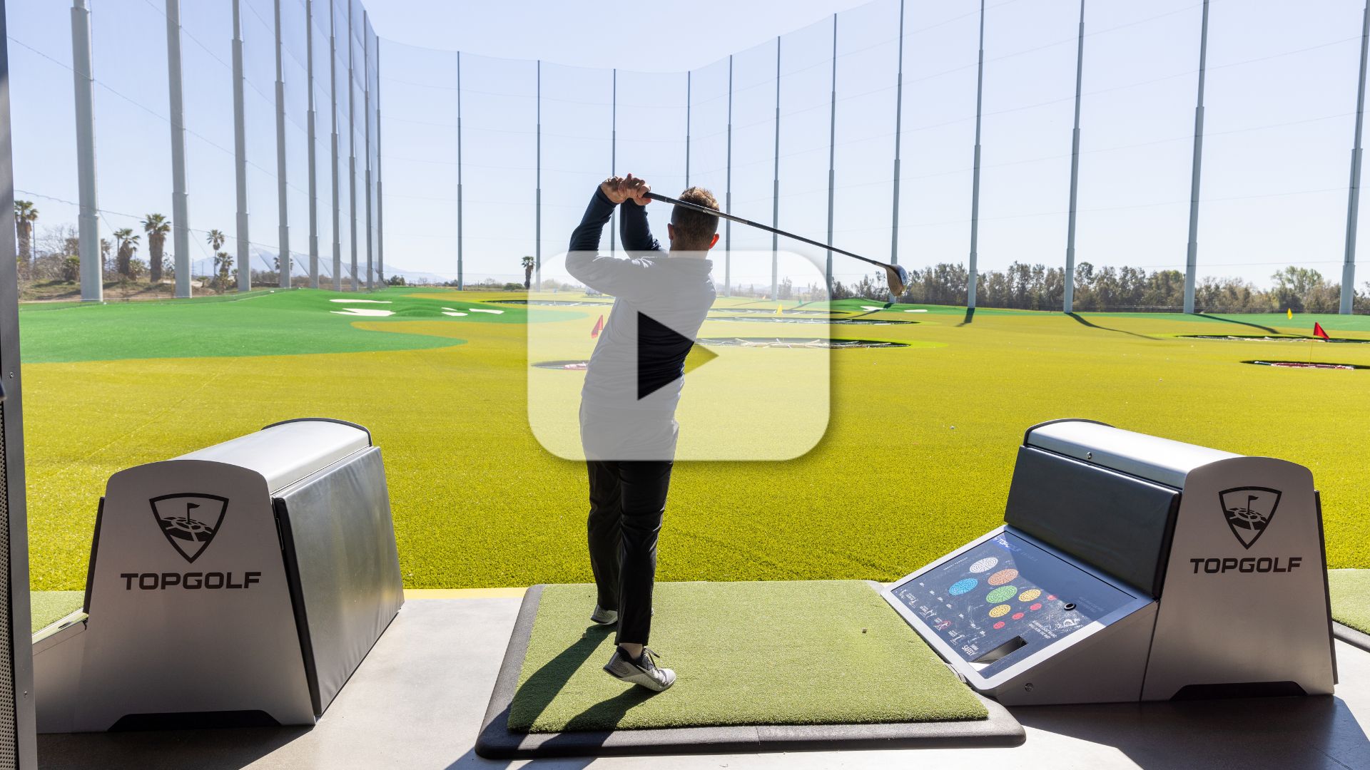 A person is taking a swing on an outdoor driving range at Topgolf. The area features green turf, targets in the distance, and a protective netting surrounding the range, and video play button overlay.