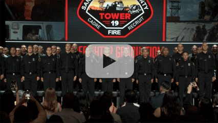 Firefighter recruits from Tower 19 stand on stage in uniform at a graduation ceremony with County Fire emblem displayed behind them.