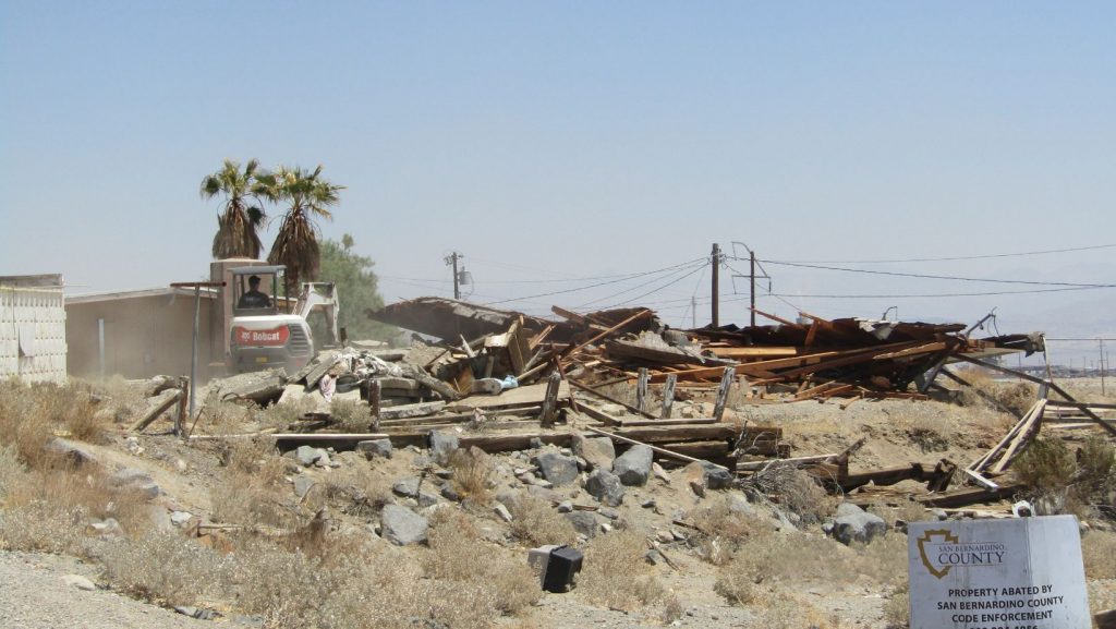 Contractor demolishes collapsed structure in Trona desert; foreground sign says property was abated by County Code Enforcement.