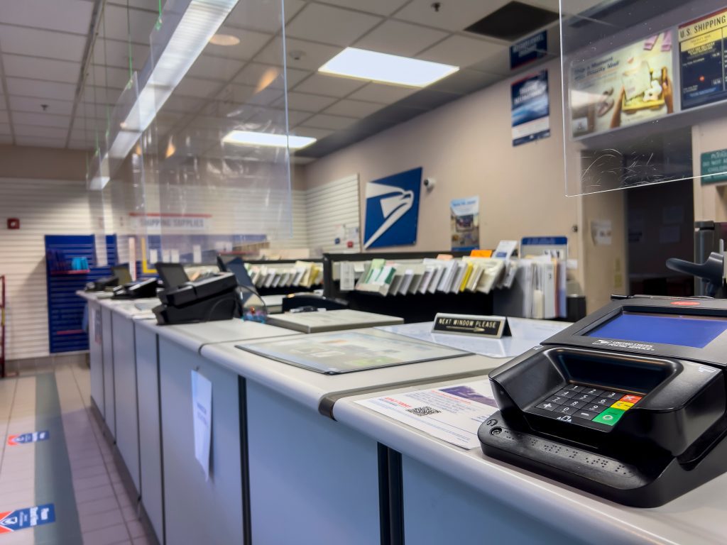 Empty post office counter with card reader, plexiglass barriers, mail slots and USPS logo on wall.
