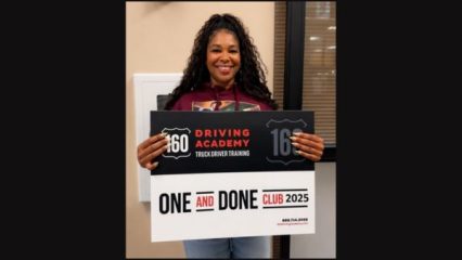 A person stands smiling and holding a sign for "160 Driving Academy Truck Driver Training" that reads "One and Done Club 2025.