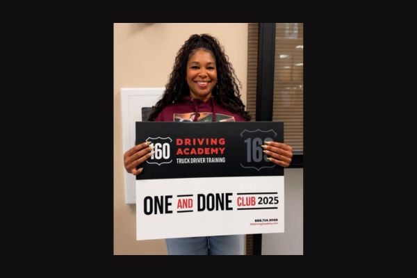 A person stands smiling and holding a sign for "160 Driving Academy Truck Driver Training" that reads "One and Done Club 2025.