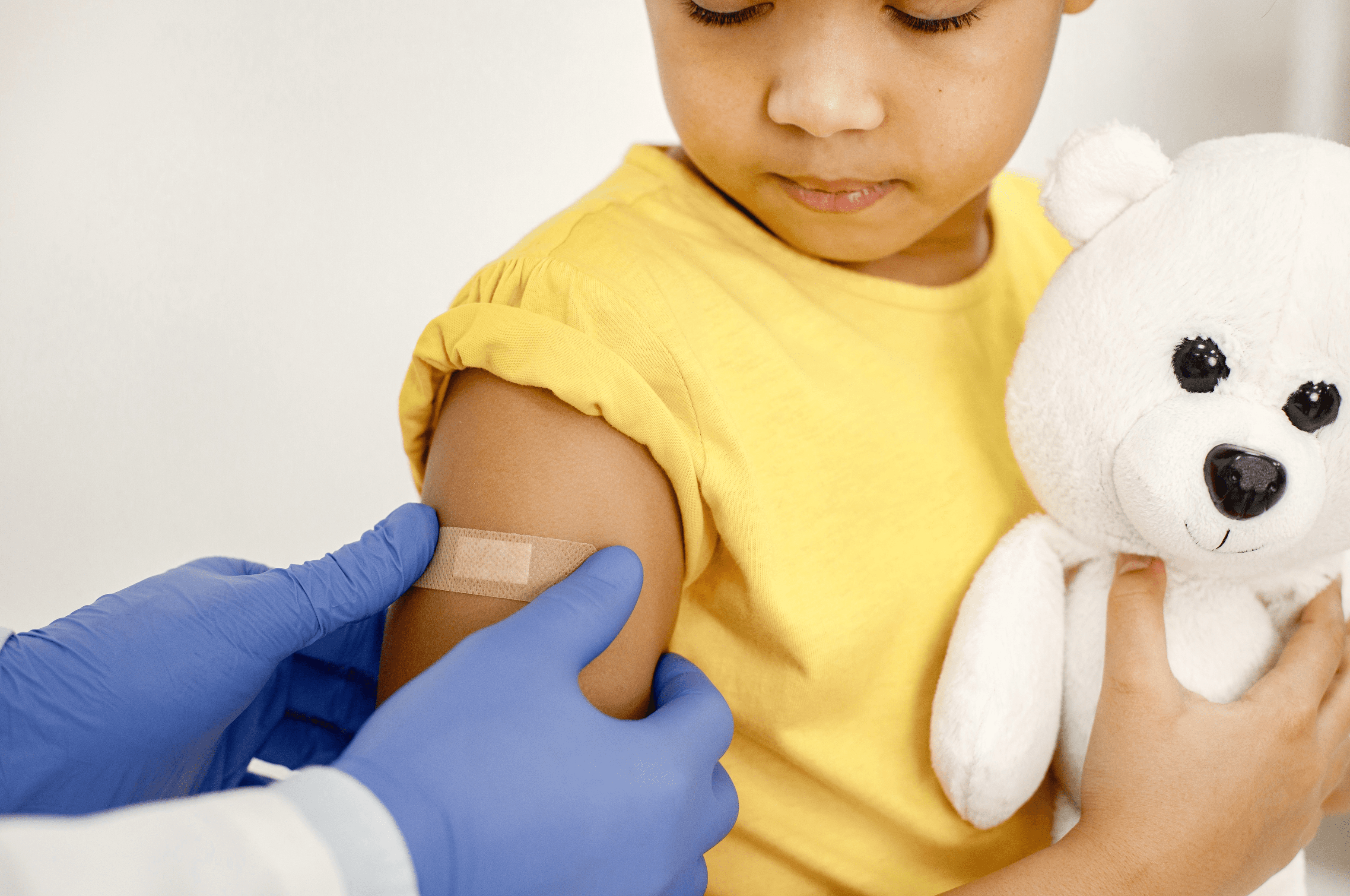 A child holding a teddy bear receives a bandage on their arm after a vaccination.