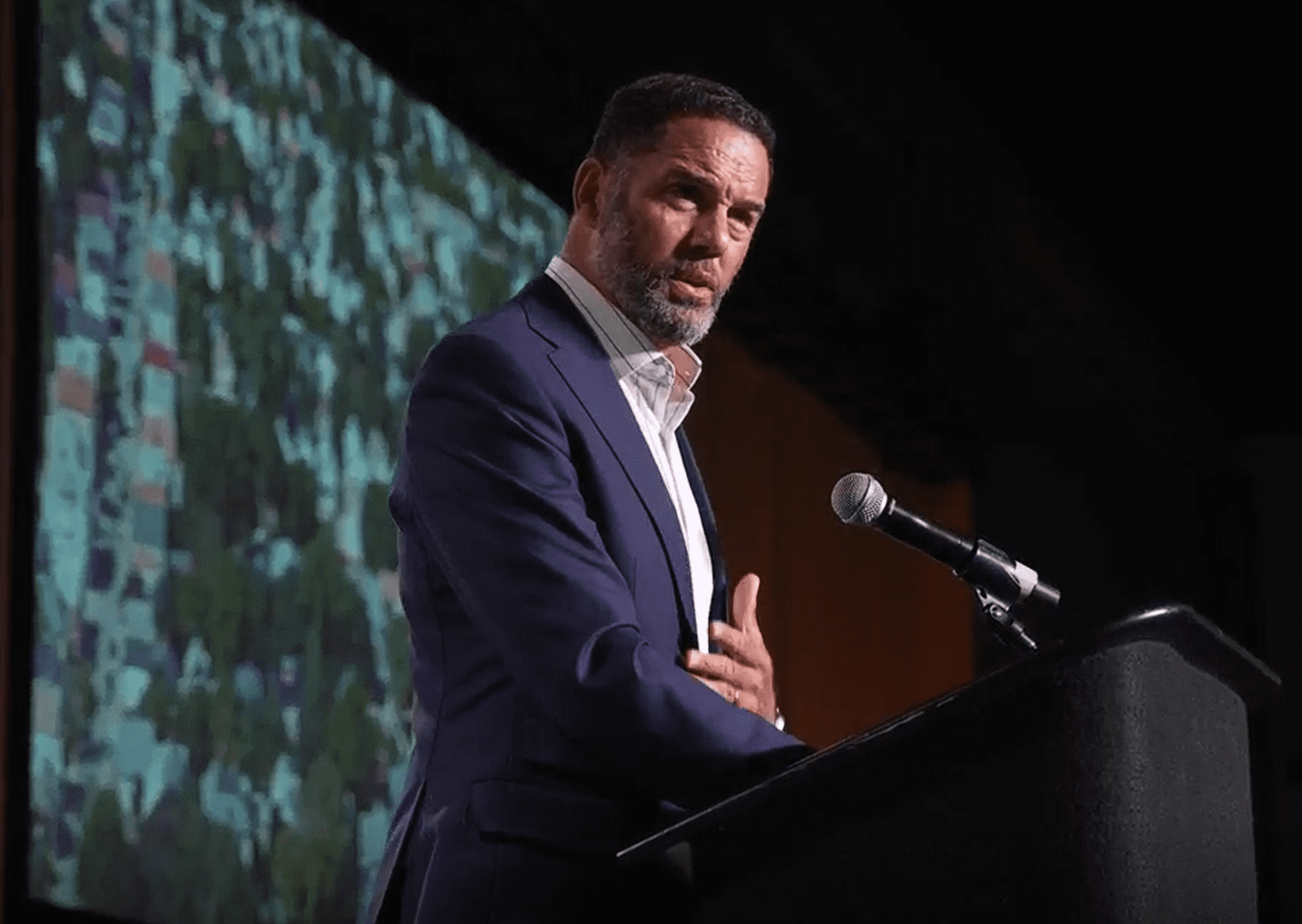 Person giving a speech at a podium with a digital screen in the background displaying abstract green visuals.