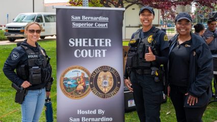 Three smiling women in uniforms stand on grass by a banner promoting the Shelter Court program