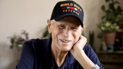 An elderly man smiles, wearing a blue shirt and a black baseball cap with World War II Veteran on it.