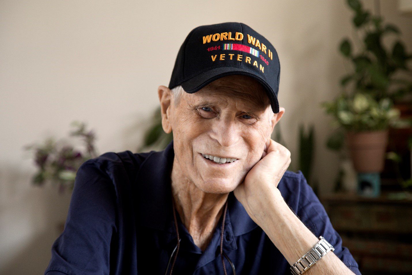 An elderly man smiles, wearing a blue shirt and a black baseball cap with World War II Veteran on it.