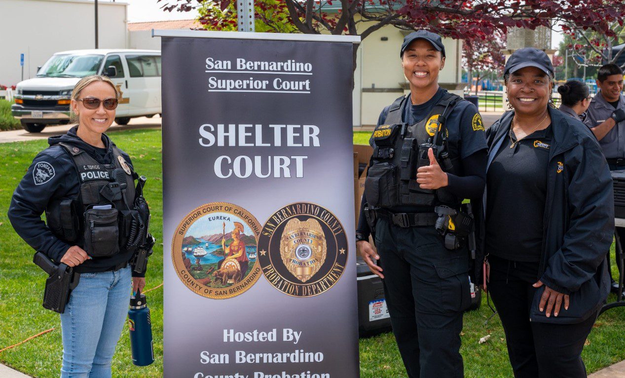 Three smiling women in uniforms stand on grass by a banner promoting the Shelter Court program