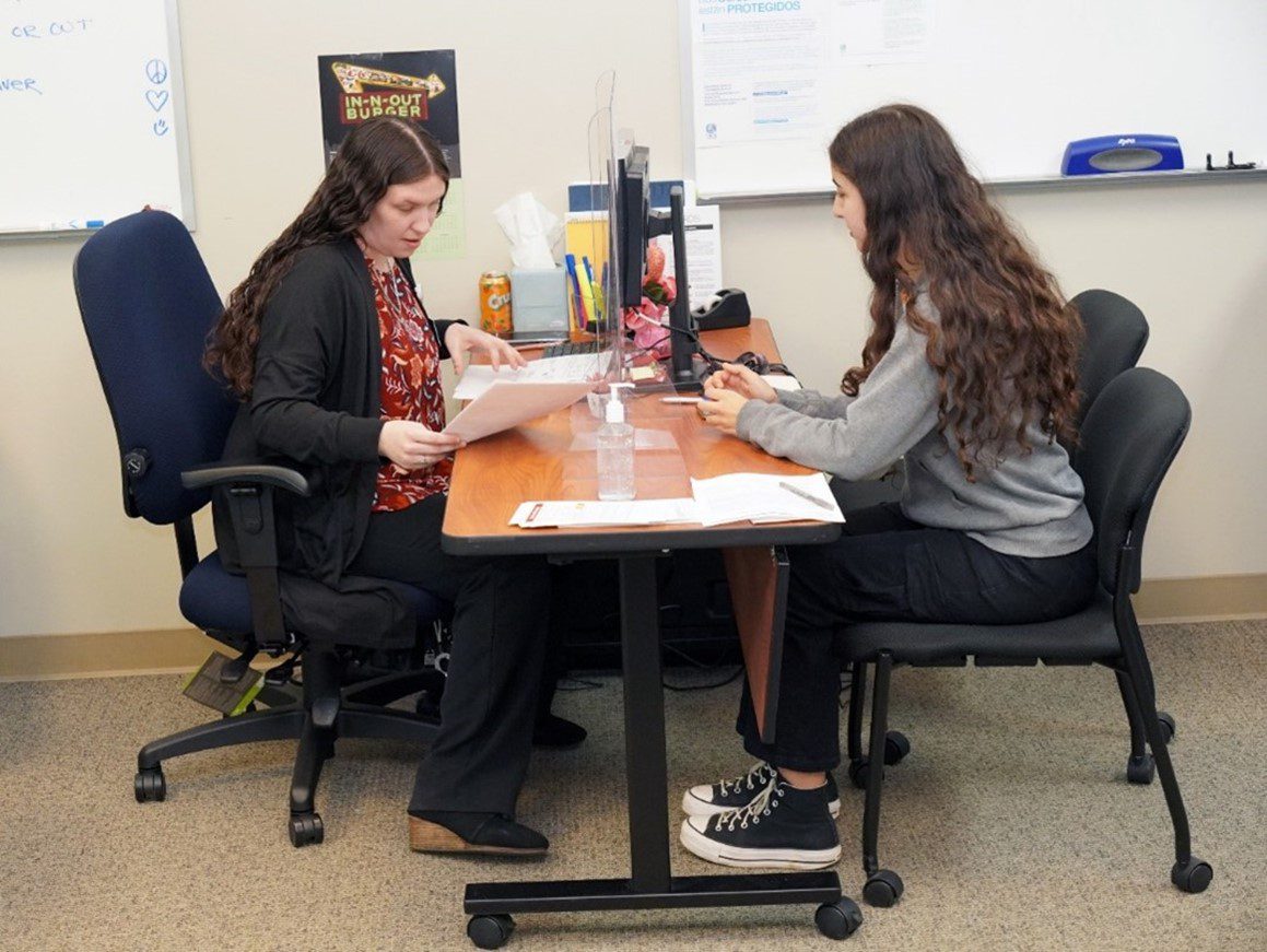 A businesswoman sitting at a desk, looking at paperwork with another woman seated across from her in an office setting.