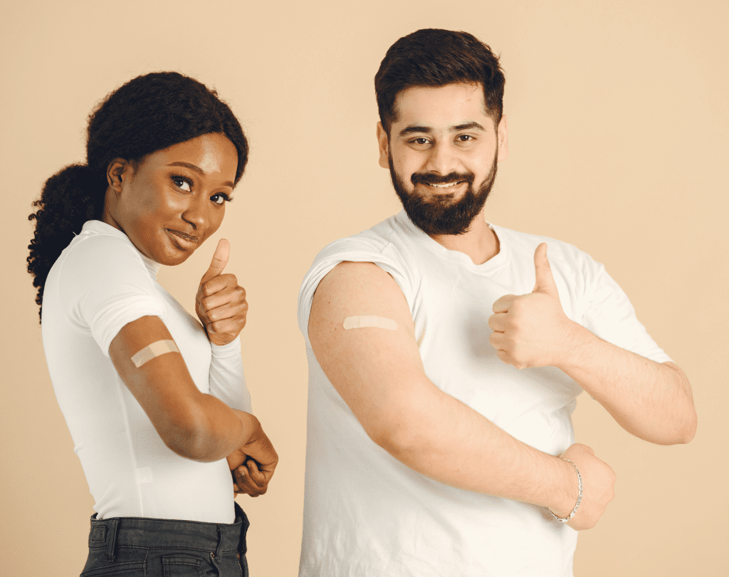 Two people smiling and giving thumbs up while showing bandages on their upper arms after getting vaccinated.