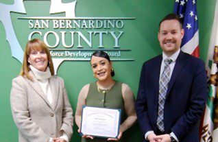 Half-photo of Three people stand before a San Bernardino County Workforce Development Board sign as a woman in green holds a certificate of achievement.