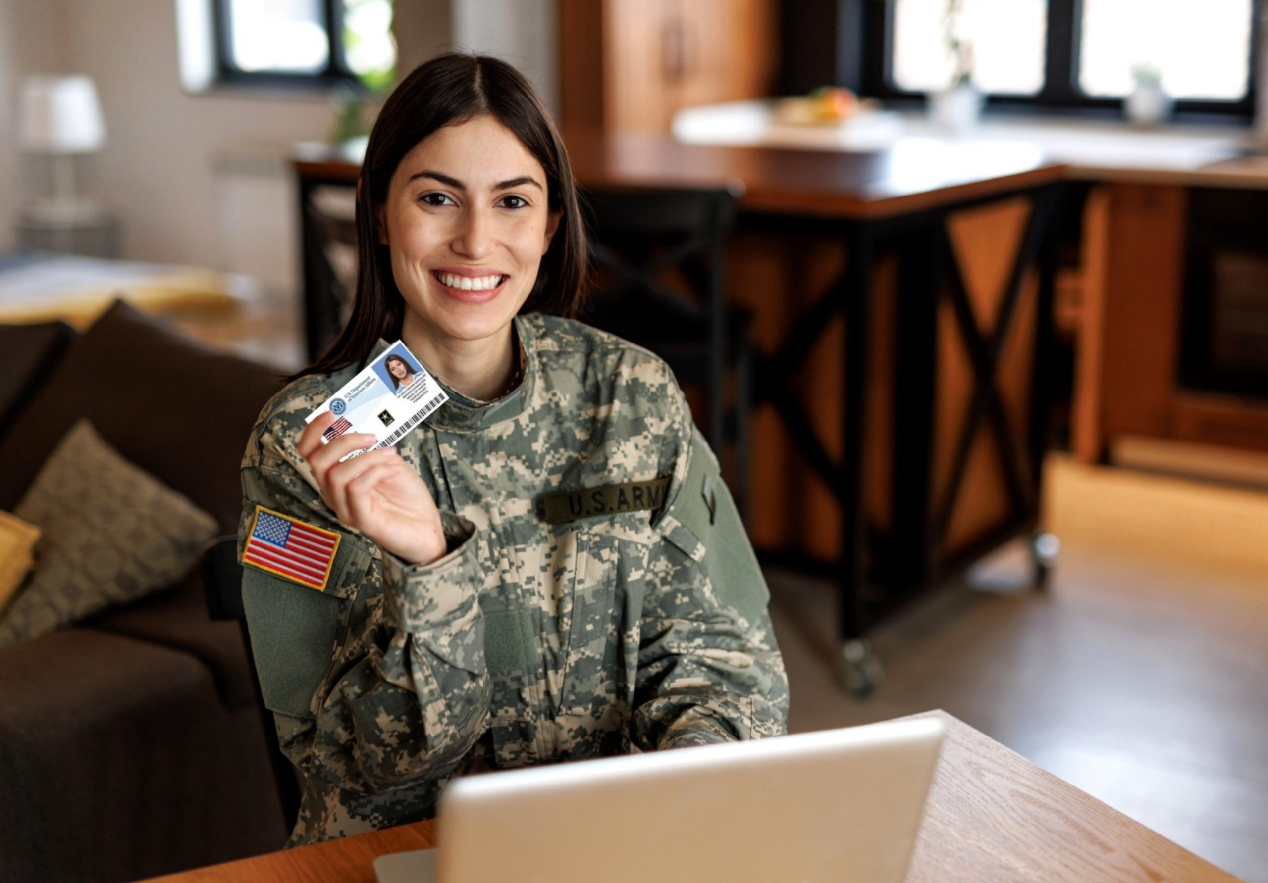 A smiling female veteran wearing a U.S. Army combat uniform and holding a U.S. Department of Veterans Affairs health identification card in a house setting.
